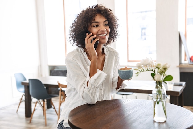 Souriante jolie jeune femme africaine se détendre à l'intérieur, à l'aide de téléphone portable, parler