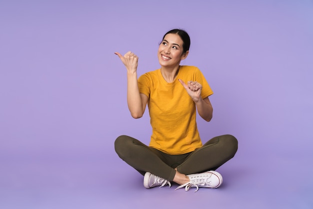 Souriante jolie jeune femme assise sur un sol avec les jambes croisées isolées sur un mur violet, pointant vers l'espace de copie