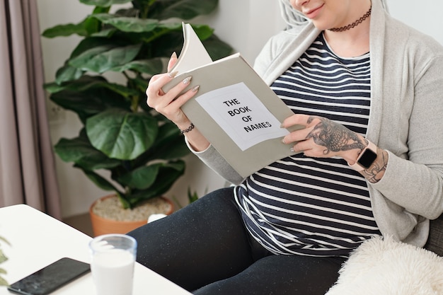 Souriante jolie jeune femme enceinte lisant un livre de noms et choisissant le nom de son enfant