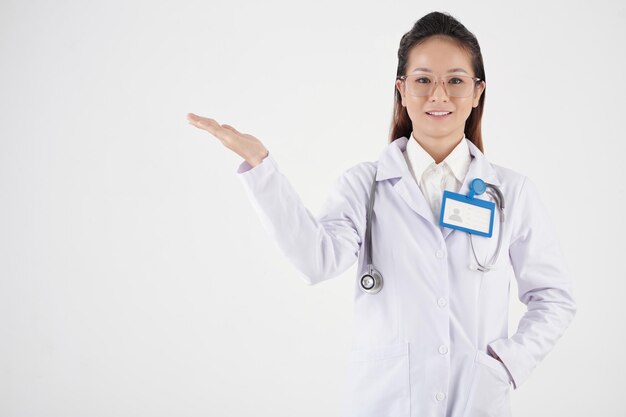 Souriante jolie jeune femme médecin avec badge sur la poitrine montrant quelque chose avec la main