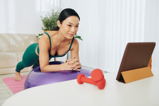 Souriante jolie jeune femme regardant un cours en ligne sur une tablette numérique et s'équilibrant en position de planche sur un ballon de fitness