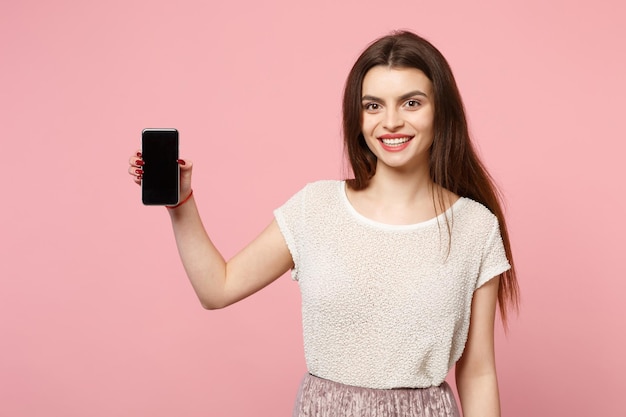 Souriante jolie jeune fille dans des vêtements légers décontractés posant isolé sur un portrait en studio de fond rose pastel. Concept de style de vie des gens. Maquette de l'espace de copie. Tenir un téléphone portable avec un écran vide vide.