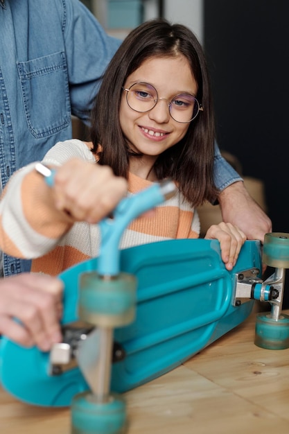 Souriante jolie jeune fille à lunettes fixant les roues du skateboard avec une clé