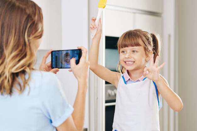 Souriante jolie petite fille posant avec une brosse en silicone et faisant un geste correct lorsque la mère la photographie dans la cuisine