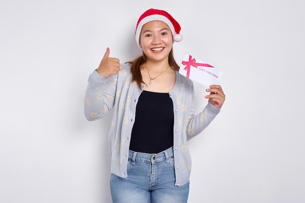 Souriante joyeuse jeune femme asiatique des années 20 dans un chapeau de père Noël tenant une carte de bon de bon de réduction de certificat-cadeau pour le magasin montrant le pouce vers le haut regardant la caméra isolée sur fond de studio blanc