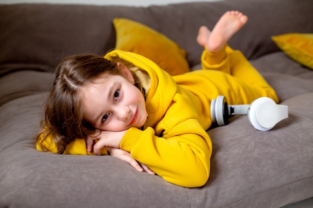 Souriante mignonne petite fille en vêtements jaunes allongée dans son lit avec un casque en écoutant de la musique