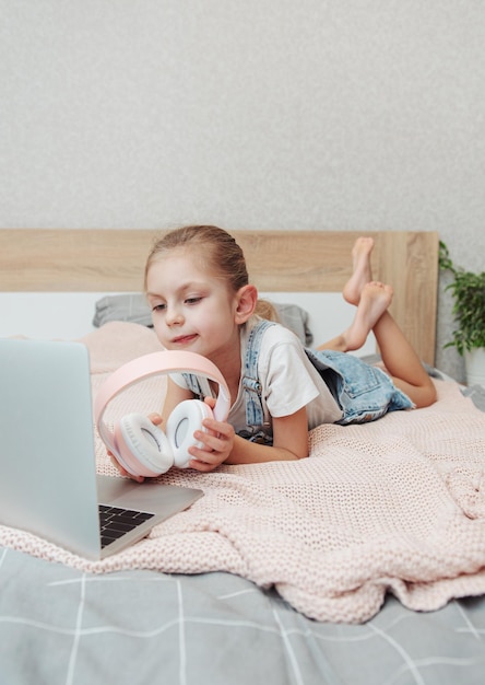 Souriante petite fille heureuse utilisant un ordinateur portable dans un casque sans fil au lit à la maison