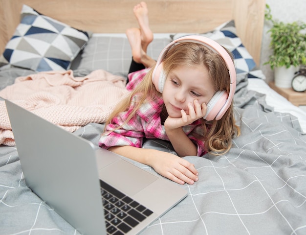 Souriante petite fille heureuse utilisant un ordinateur portable dans un casque sans fil au lit à la maison