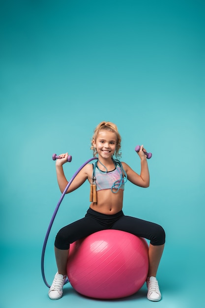 Souriante petite fille sportive faisant des exercices avec différents équipements sportifs isolés sur mur bleu