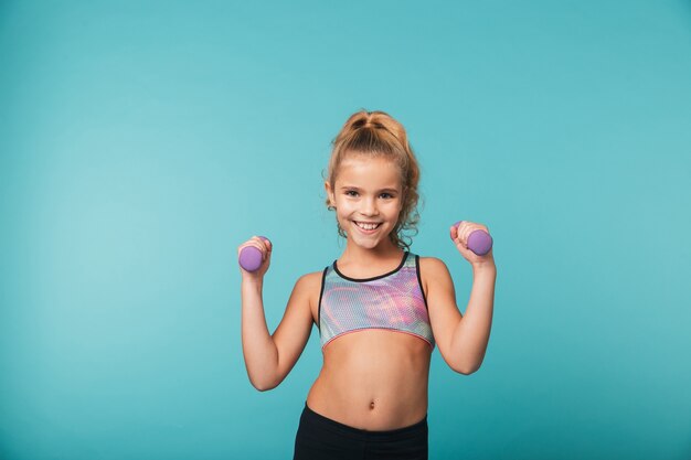 Souriante petite fille sportive faisant des exercices avec des haltères isolés sur mur bleu