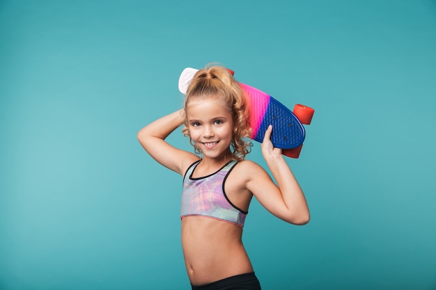 Souriante petite fille sportive jouant avec une planche à roulettes isolée sur mur bleu