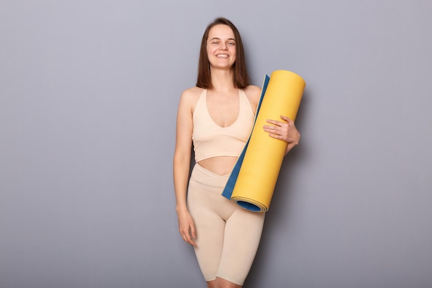 Souriante sportive gaie tenant un tapis de yoga isolé sur fond gris faisant des exercices de sport regardant la caméra avec un visage heureux mode de vie sain