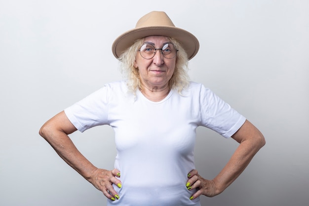 Souriante vieille femme dans un chapeau tenant les mains sur une ceinture sur un fond clair.