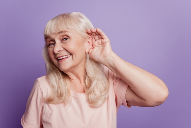 Souriante vieille femme avec la main sur l'oreille écouter des sons isolés sur fond violet