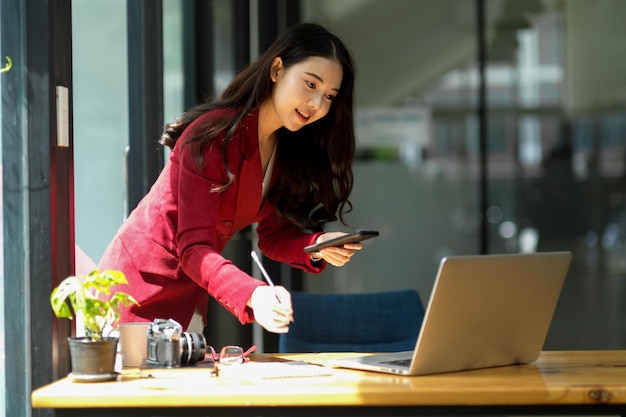 Souriez une jeune femme d'affaires asiatique utilisant un smartphone tout en travaillant sur un ordinateur portable et prenez des notes dans son journal personnel au bureau.