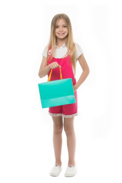 Sourire accro du shopping petite fille avec panier isolé sur blanc. Une fille heureuse tient un sac en papier. Enfance. Enfant shopper en combinaison de mode. Préparation et célébration des vacances. Achats et vente.