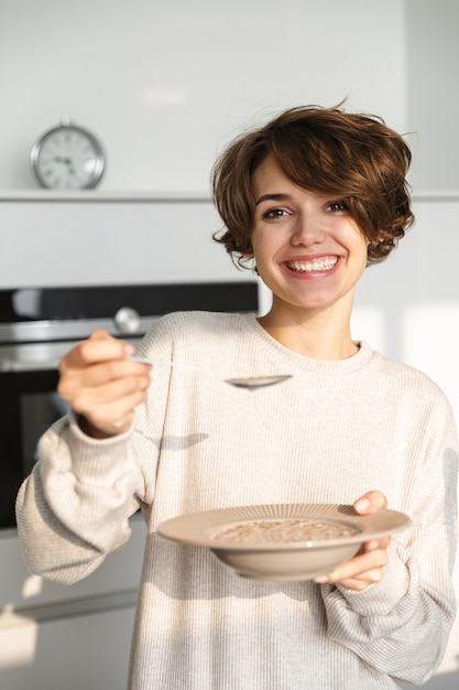 Sourire affamé jeune femme debout au réfrigérateur, manger du porridge