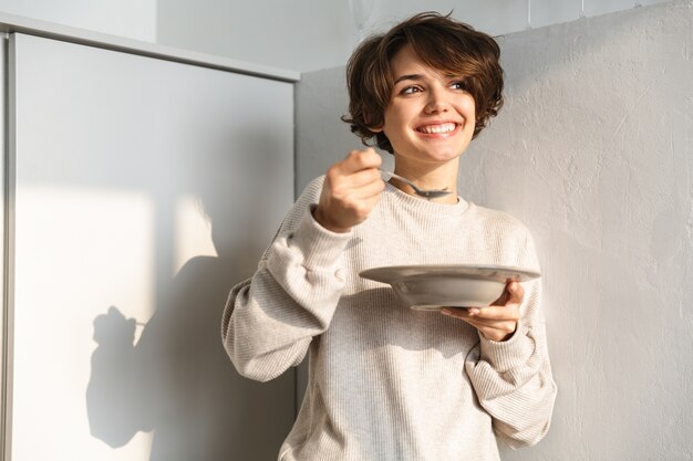 Sourire affamé jeune femme debout au réfrigérateur, manger du porridge