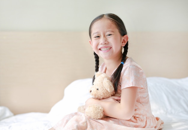 Sourire asiatique petite fille câlin nounours assis sur le lit à la maison