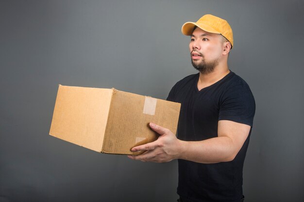 Sourire beau livreur asiatique portant casquette, donnant et transportant des colis, boîte en carton, journée de déménagement et concept de livraison express