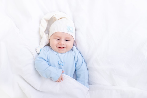 Sourire bébé garçon sur le lit sous la couverture dans un bonnet