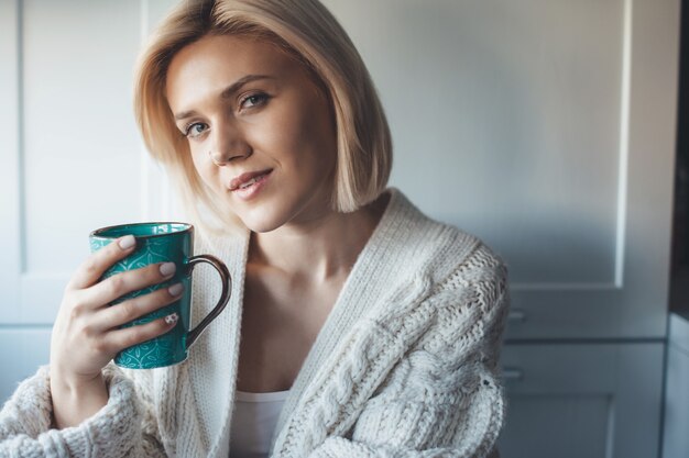 Sourire belle femme aux cheveux blonds, boire une tasse de thé dans la cuisine portant un pull en tricot