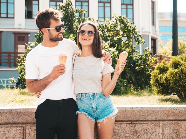 Photo sourire belle femme et son beau petit ami femme en vêtements d'été décontractés bonne famille joyeuse couple posant sur le fond de la rue à lunettes de soleil manger de la crème glacée savoureuse dans un cône de gaufres