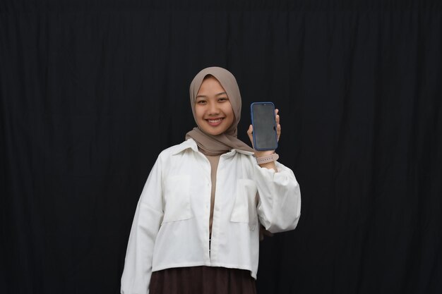 Photo sourire belle fille asiatique en hijab, montrant un écran vertical de téléphone portable vierge