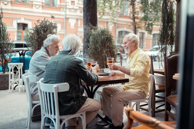 Sourire et boire. Hommes aux cheveux gris à la retraite souriant et buvant de l'alcool assis à l'extérieur du pub