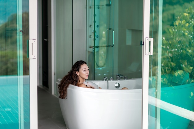 Sourire brune posant en position allongée dans un bain de mousse près d'une fenêtre ouverte