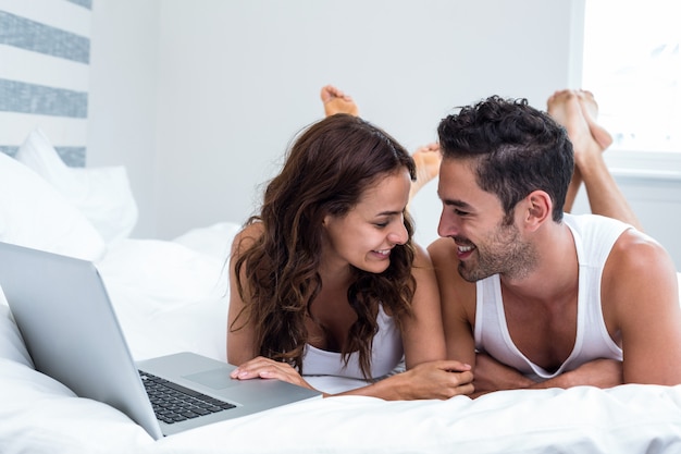 Sourire couple avec ordinateur portable en position couchée sous couverture