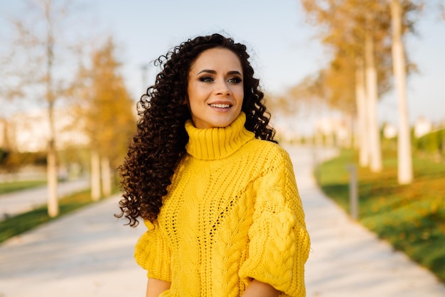 Sourire doux sourire fille dans un chandail brillant avec des boucles de cheveux noirs posant dans le parc en automne