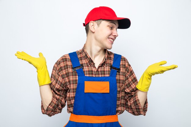 Sourire écartant les mains jeune homme de nettoyage portant un uniforme et une casquette avec des gants