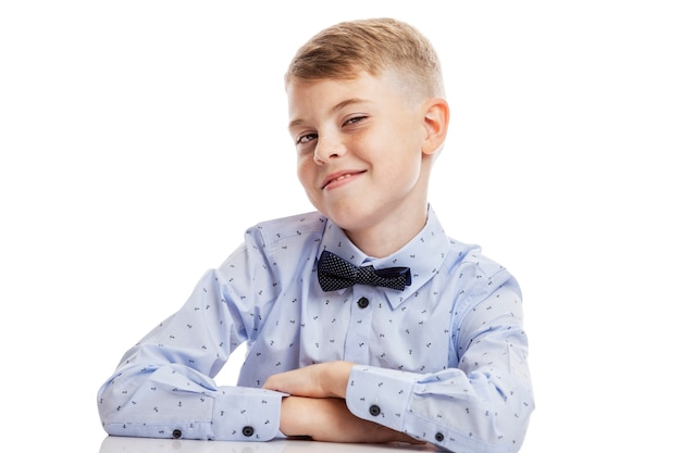 Sourire écolier dans une chemise bleue avec un nœud papillon est assis à la table.