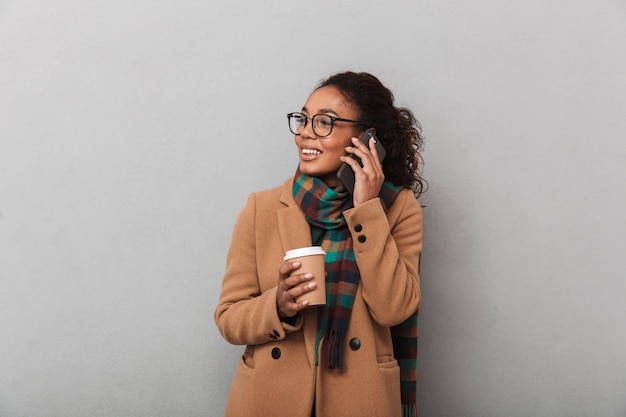 Sourire femme africaine portant manteau debout, parler au téléphone mobile, tenant une tasse de café