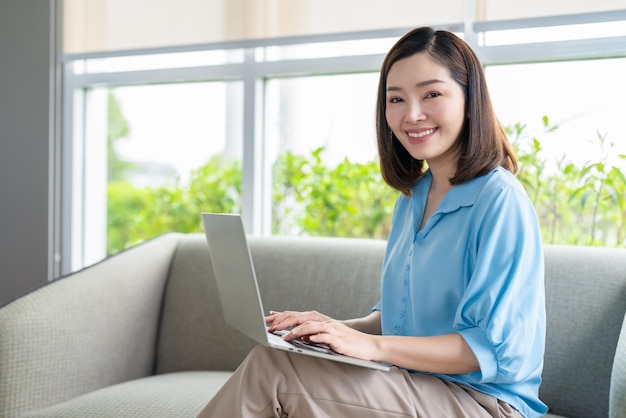 Sourire de femme asiatique portant une chemise bleue à l'aide d'un ordinateur portable et assis sur un canapé dans le salon à la maison