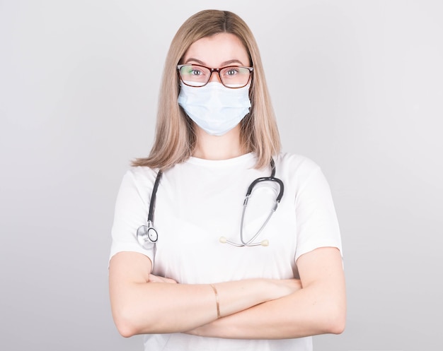 Sourire de femme médecin en masque médical, lunettes, avec les bras croisés sur fond blanc
