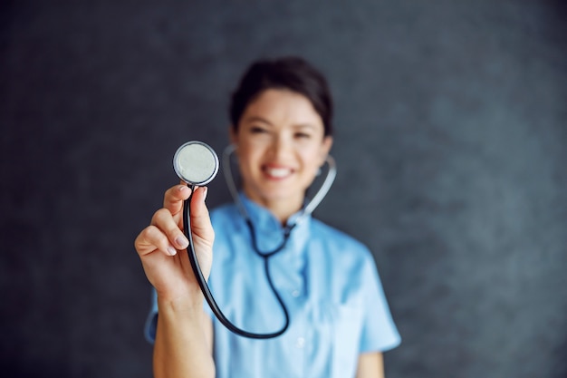 Sourire femme médecin tenant le stéthoscope vers la caméra comme elle examine les poumons. Mise au point sélective sur le stéthoscope.