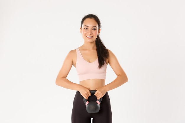 Sourire fille asiatique forte et mince de remise en forme, musculation à la maison, tenant un équipement d'entraînement, faire des squats avec exercice kettlebell, fond blanc.