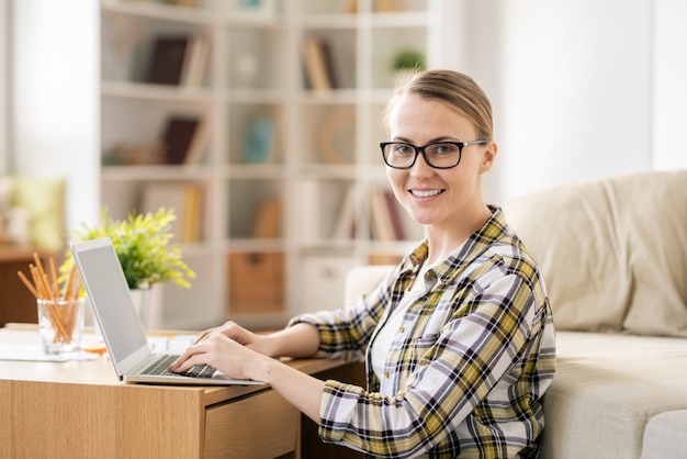 Sourire fille moderne dans des verres assis dans le salon tout en utilisant un ordinateur portable et obtenir des connaissances via Internet