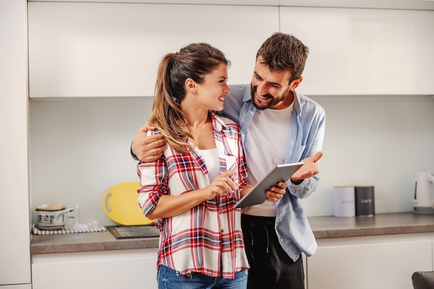 Sourire heureux couple debout ensemble dans la cuisine et à l'aide de tablette pour rechercher une recette