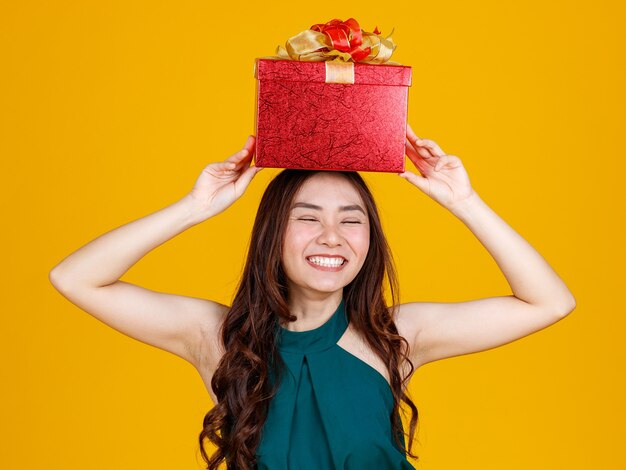 Sourire heureux face à une jolie fille asiatique aux cheveux noirs tenant une boîte-cadeau au-dessus de la tête avec un délicieux et excité, tourné en studio sur fond jaune. Célébrer et festival concept.