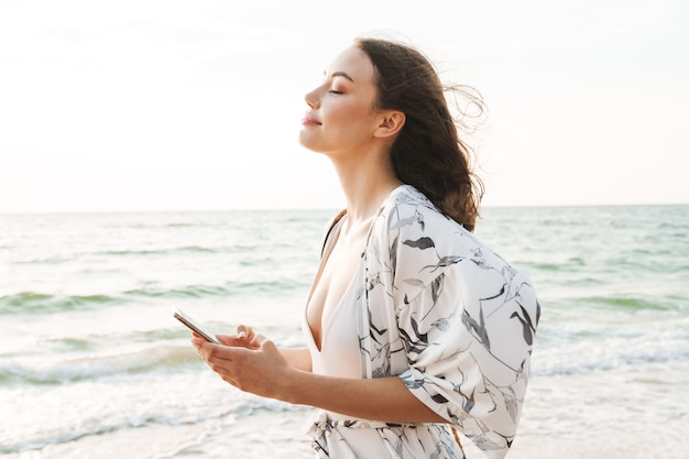 sourire heureux heureux jeune belle femme à la plage marchant dans une belle matinée ensoleillée à l'aide d'un téléphone portable.