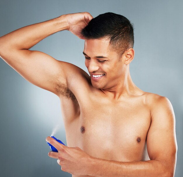 Photo sourire heureux et homme avec un spray déodorant pour l'hygiène parfum frais et parfum après une douche bonheur propre et gars pulvérisant son aisselle pour éviter les odeurs ou les odeurs isolées par un fond gris