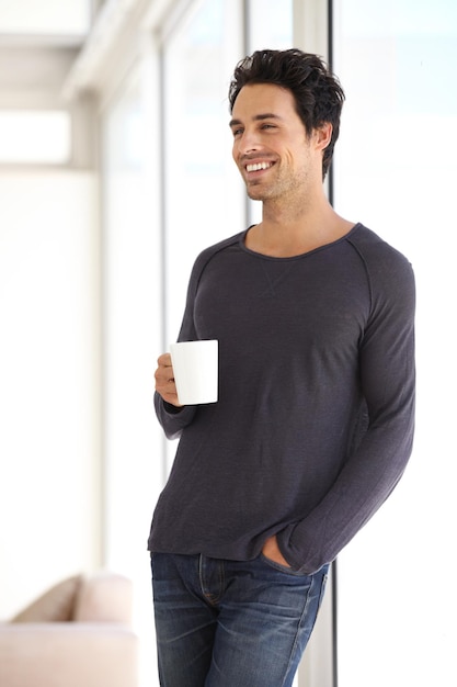 Sourire heureux et homme avec une tasse de café debout près de la fenêtre dans une maison de vacances Calme relaxant et séduisant homme buvant un café au lait dans une tasse dans le salon en pensant ou en rêvant