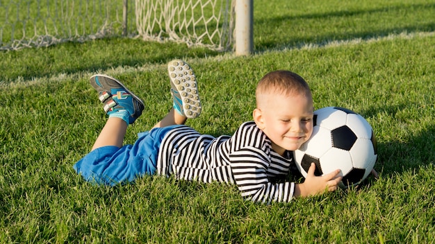 Sourire heureux jeune garçon allongé sur l'herbe verte sur un terrain de sport dans la lumière du soir avec un ballon de foot