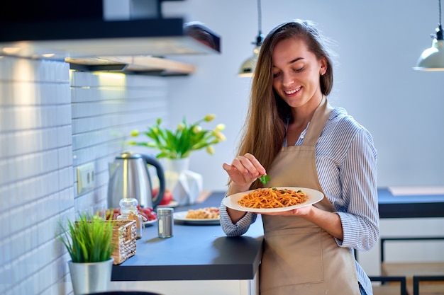 Sourire heureux joyeux mignon femme de cuisine portant un tablier décore une nourriture fraîche avec des feuilles de persil pour le dîner