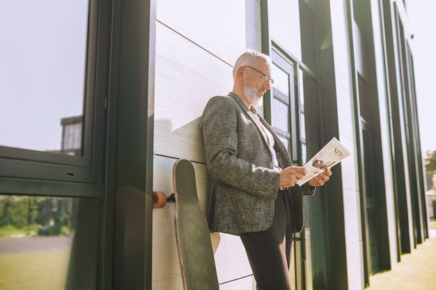 Sourire heureux à lunettes homme mûr s'appuya contre le mur du bâtiment en lisant un journal à l'extérieur