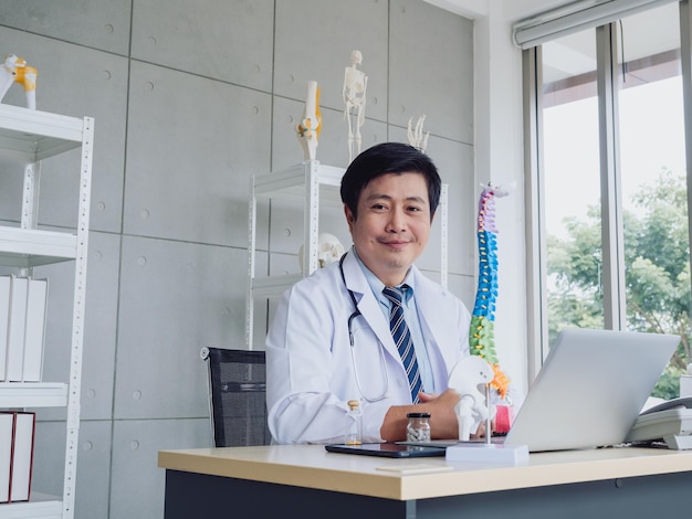 Sourire heureux Médecin orthopédique masculin adulte asiatique en costume blanc travaillant avec un ordinateur portable et une tablette numérique modèle de colonne vertébrale colorée et une bouteille de suppléments de vitamines sur le bureau près de la fenêtre en verre