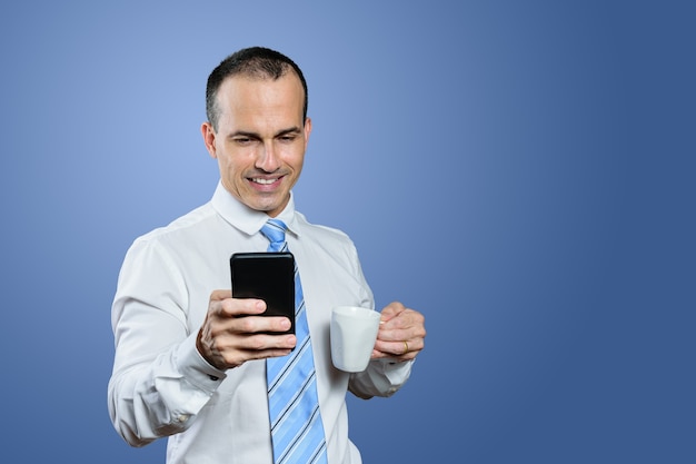 Sourire homme brésilien mature dans des vêtements formels tenant un smartphone et une tasse. Fond bleu.